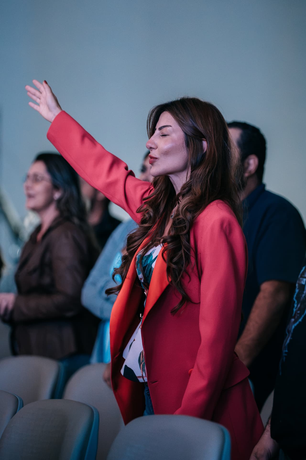 Mulher de blazer vermelho com a mão levantada em adoração durante um culto da Celeiro.